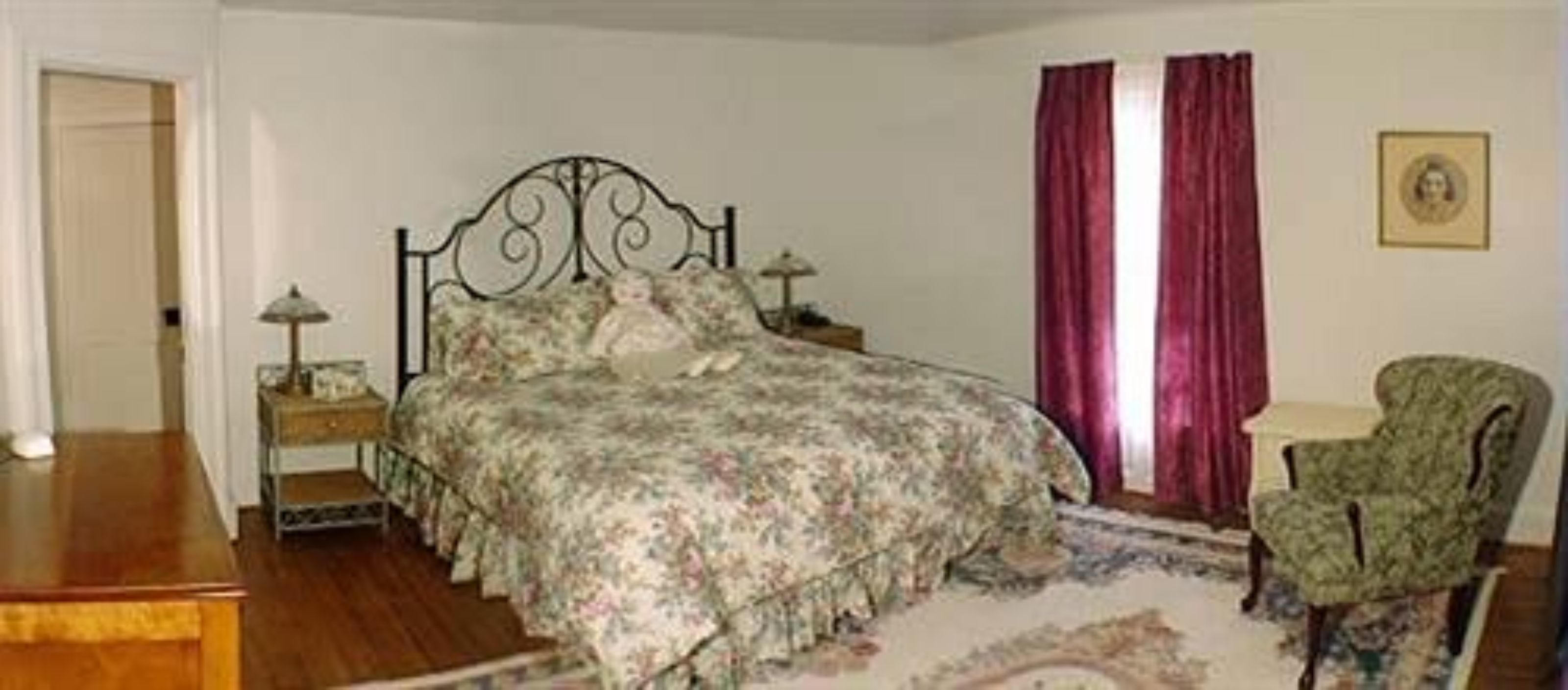 A bedroom with a floral bedspread, a metal bed frame, a nightstand with a lamp, a chair, and a window with red curtains.
