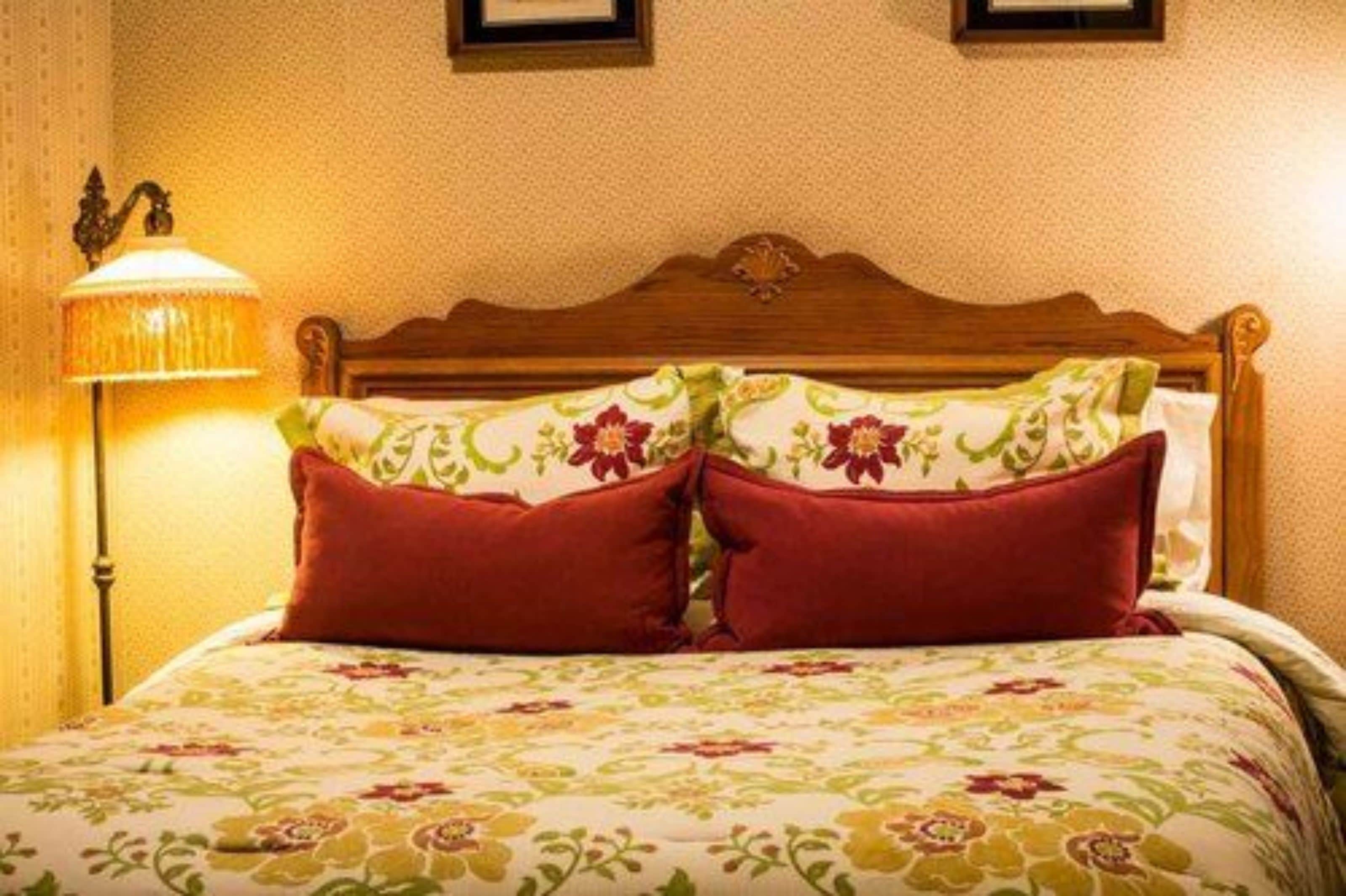 A neatly made bed with floral bedding and two red pillows, a wooden headboard, and a wall-mounted lamp.