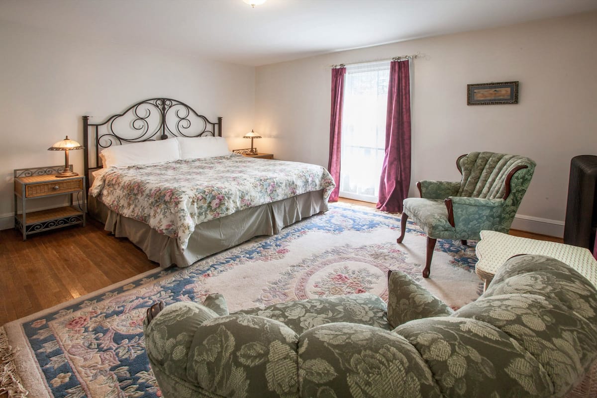 Une chambre à coucher avec un lit, deux tables de chevet avec des lampes, un fauteuil vert, un tapis à motifs et une fenêtre avec des rideaux.