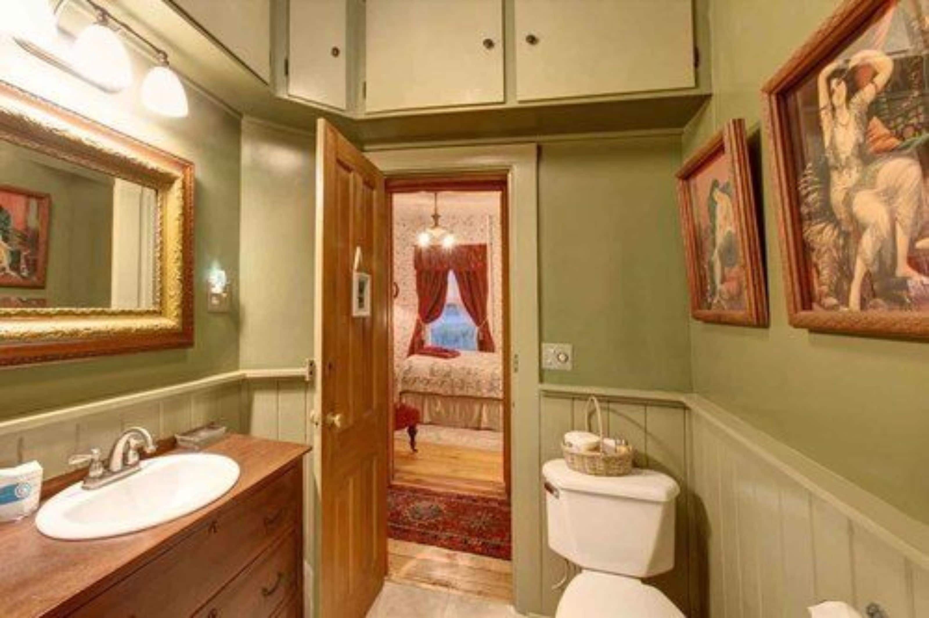 A bathroom with a wooden vanity, a white sink, a toilet, and a door leading to a bedroom with a bed and a red curtain.