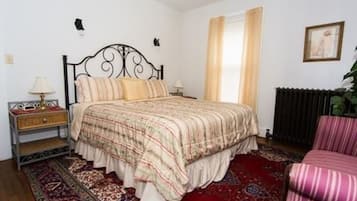 Ein Schlafzimmer mit Bett, Nachttisch, Stuhl, Heizkörper und einem Fenster mit Vorhängen.
