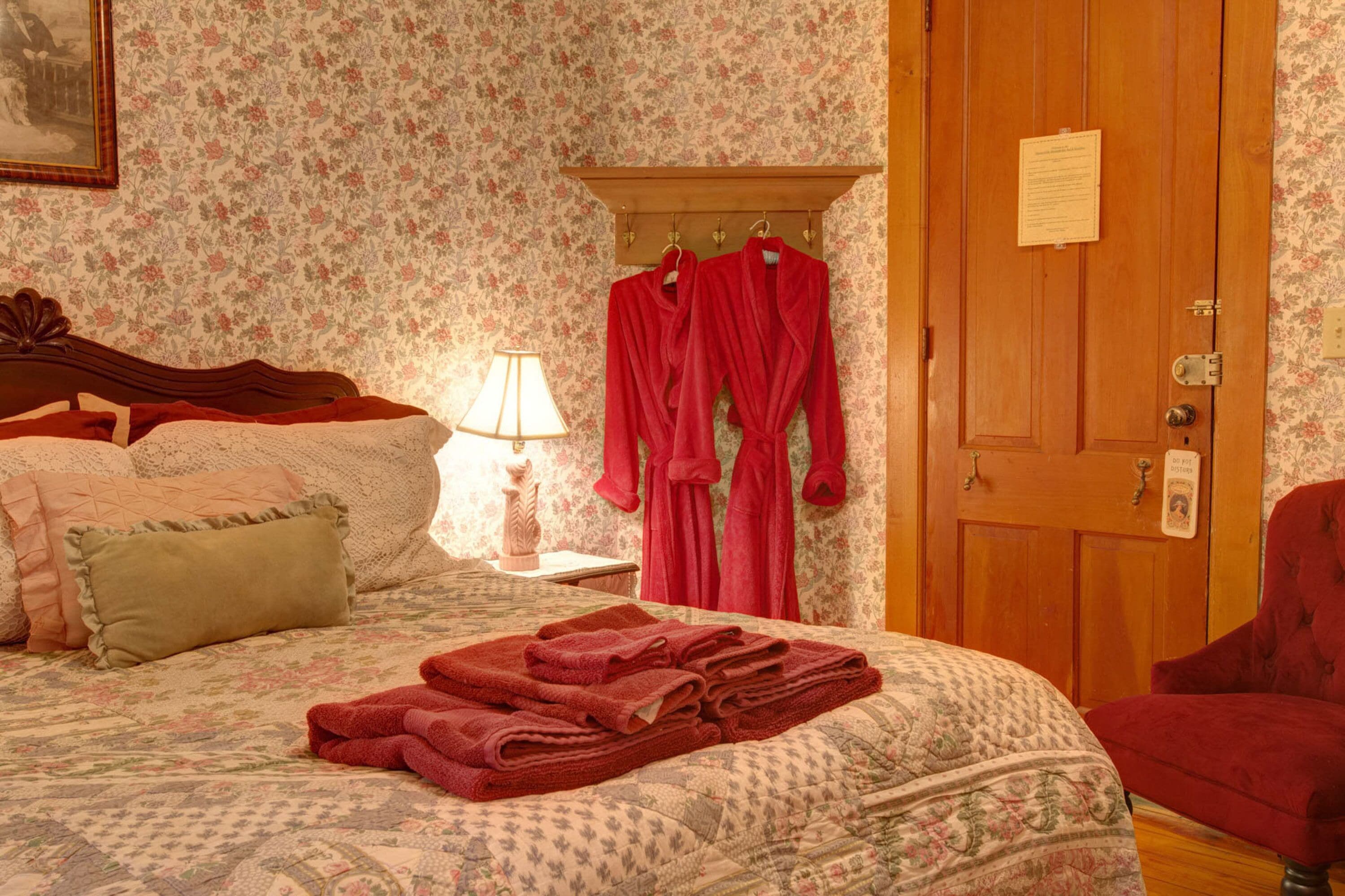 A hotel room with floral wallpaper, a wooden bed with a quilt, a nightstand with a lamp, and a red chair.