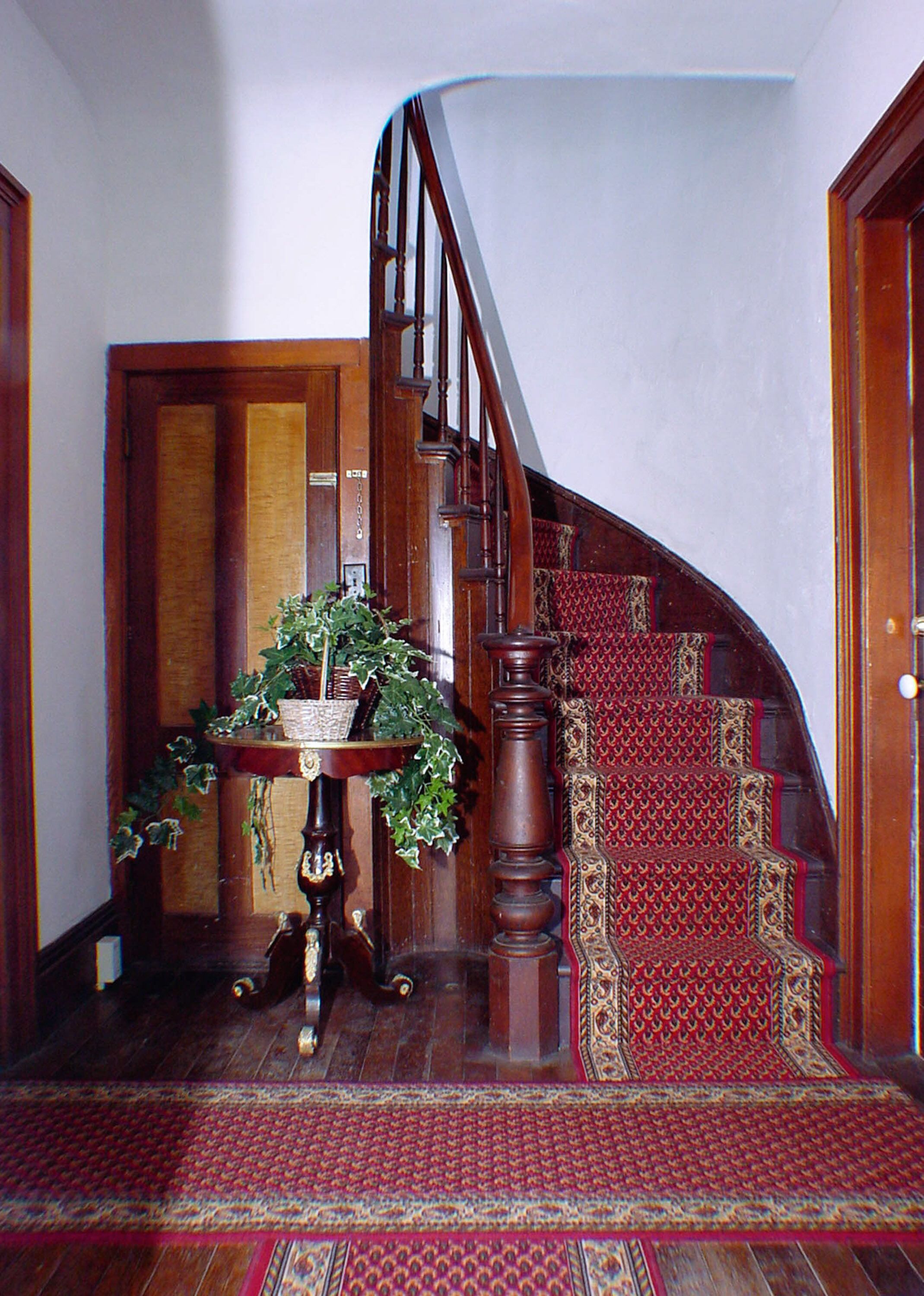 A wooden staircase with a red carpet, a wooden door, and a table with a plant.