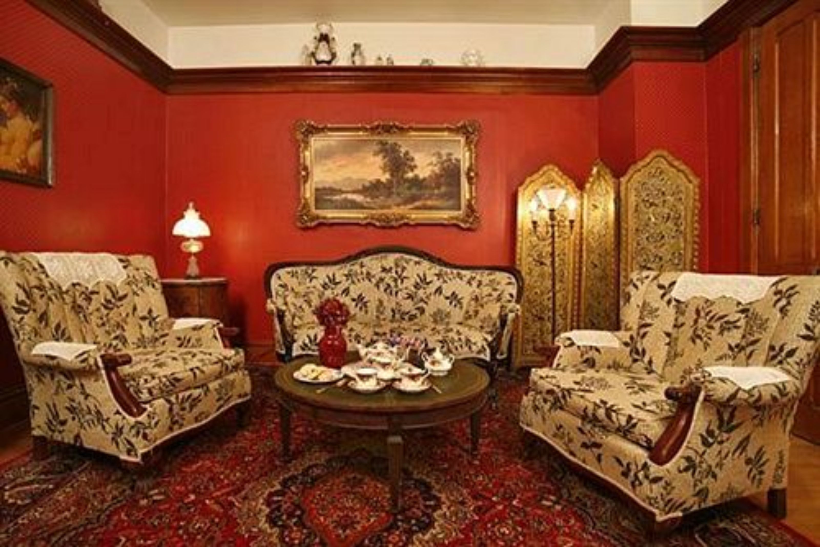 A room with a red wall, two floral armchairs, a round table with tea sets, and a framed landscape painting.