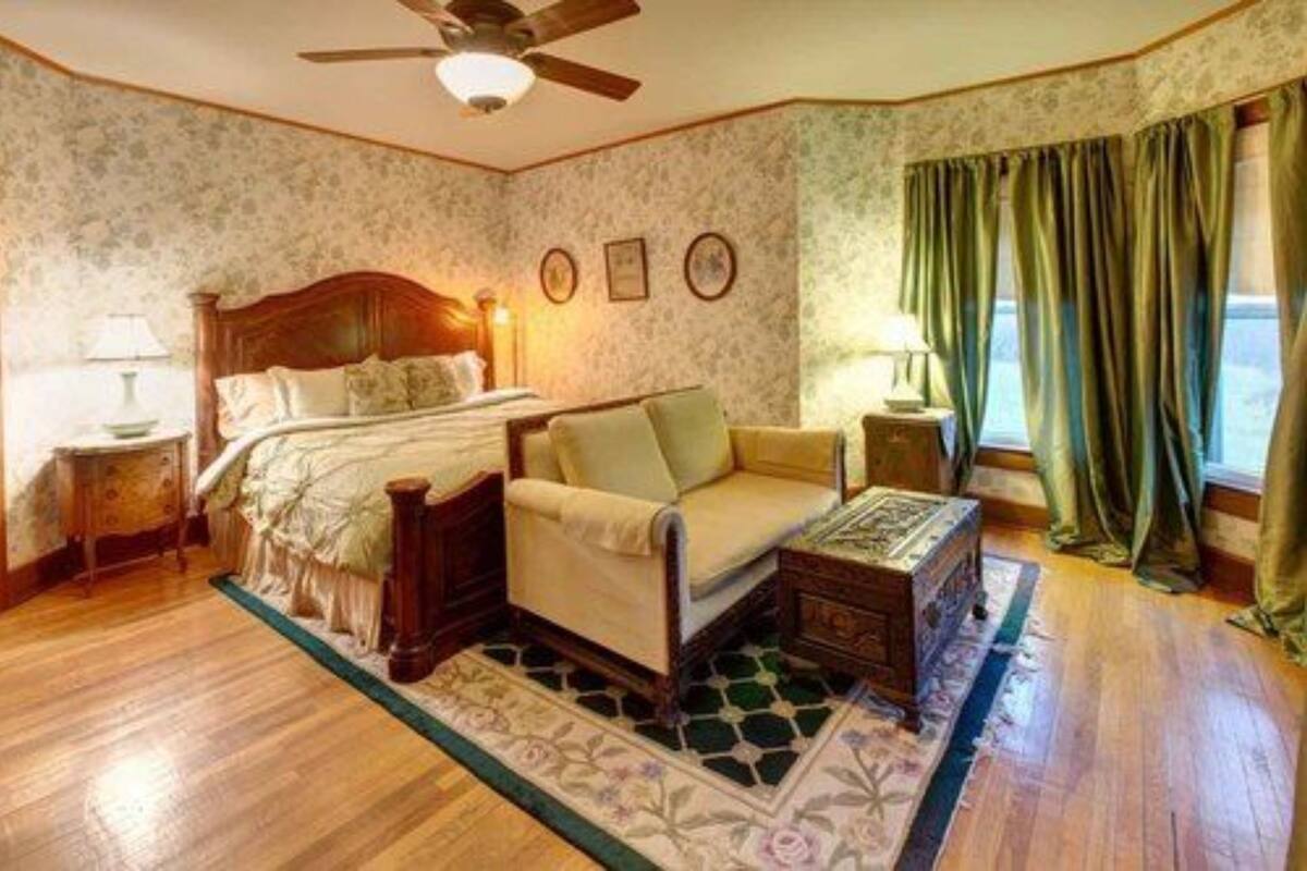 Une chambre à coucher avec un lit en bois, une chaise, une table de chevet, une commode et une fenêtre avec des rideaux.