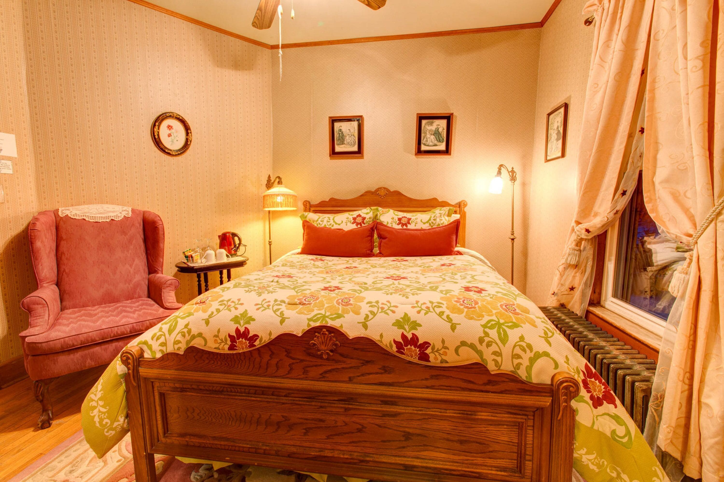 Un dormitorio con una cama de madera, un sillón rosa, una mesita de noche con una lámpara y cuadros enmarcados en la pared.
