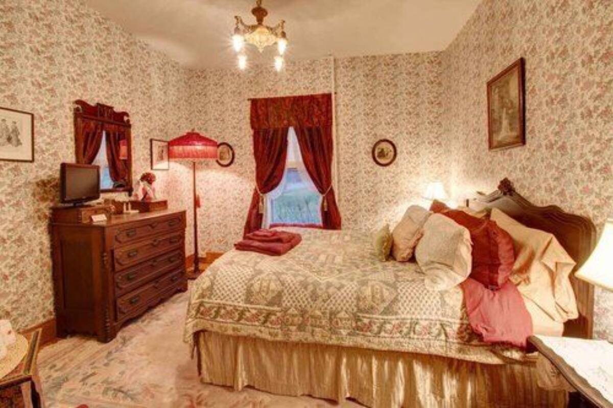 Une chambre avec un papier peint à motifs, une commode en bois, un lit avec de nombreux coussins, une lampe rouge et un lustre.
