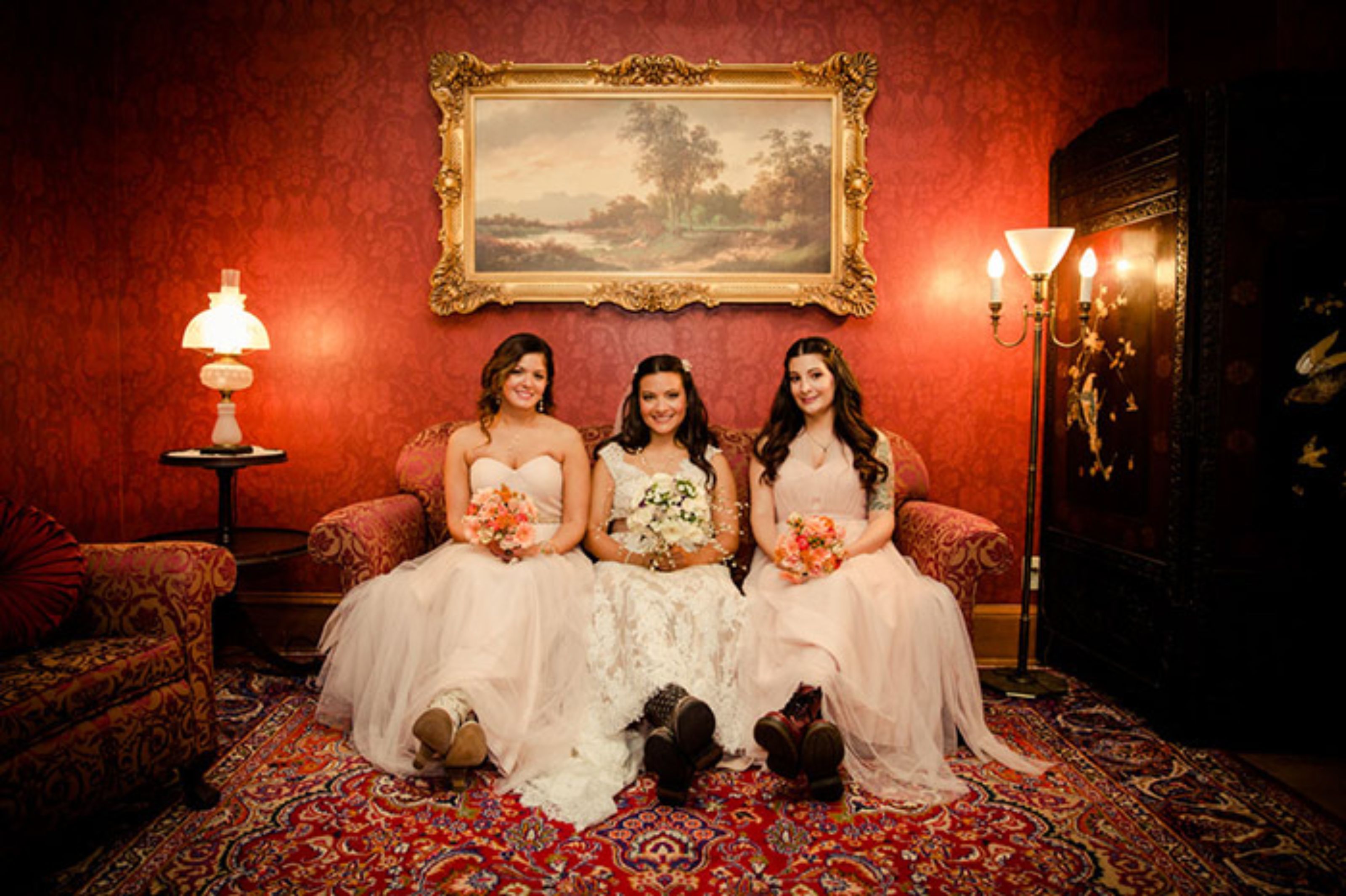 Trois femmes, vêtues de robes de mariée, sont assises sur un canapé à motifs dans une pièce aux murs rouges, où l’on aperçoit un tableau encadré et un lampadaire.