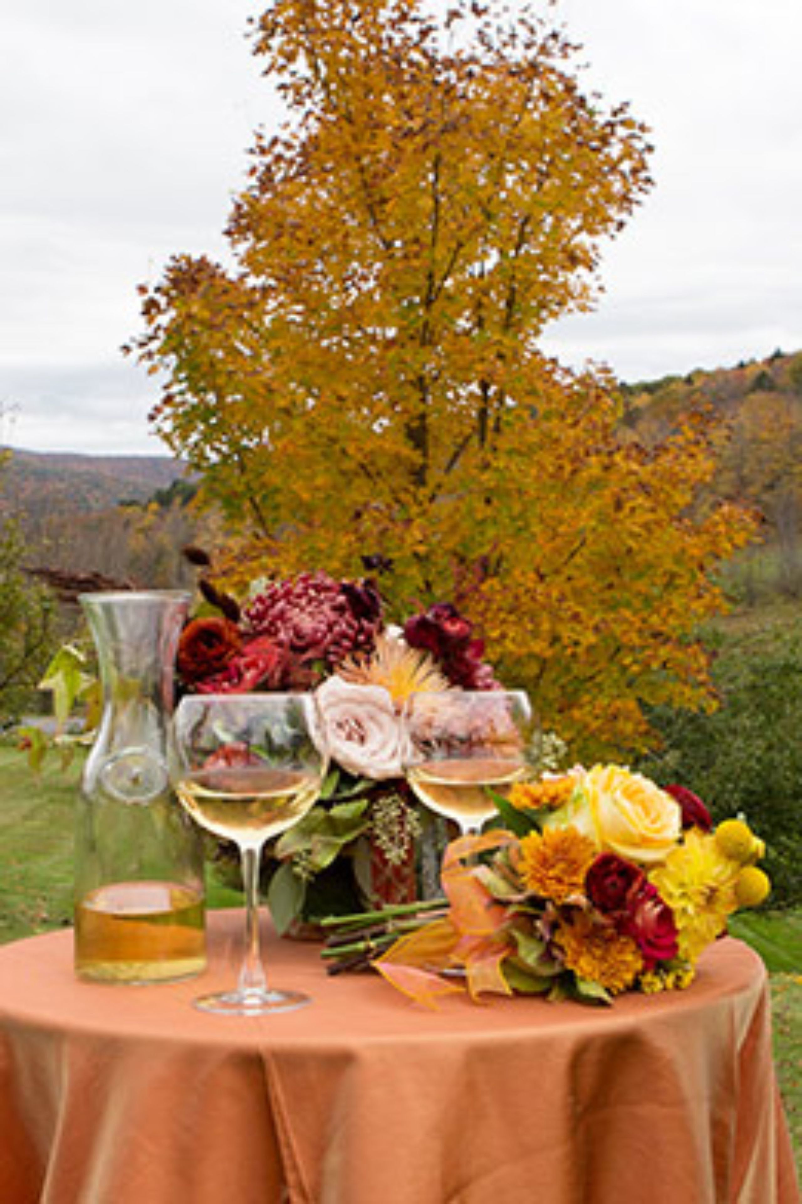 Una mesa al aire libre con mantel, un florero de vidrio con flores y una copa de vino blanco.