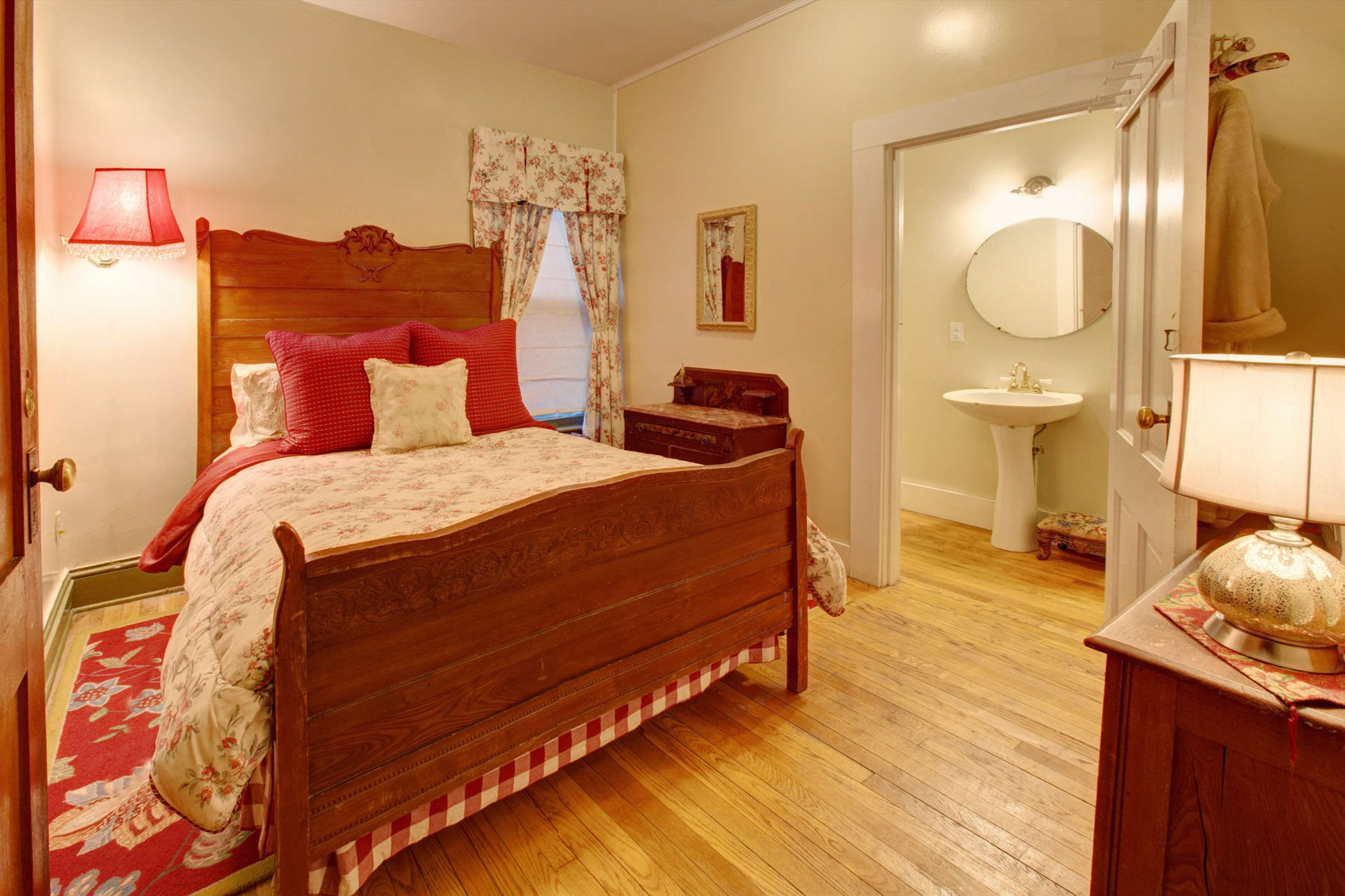 Ein Schlafzimmer mit einem hölzernen Bett, einem Nachttisch, einer Nachttischlampe, einer roten Lampe, einer zudeckenden Bettdecke mit Blumenmuster, einem rot-weißen Schachbrettteppich, einem Holzboden, einem Badezimmer mit Waschbecken, einem Spiegel und einem Handtuchhalter.