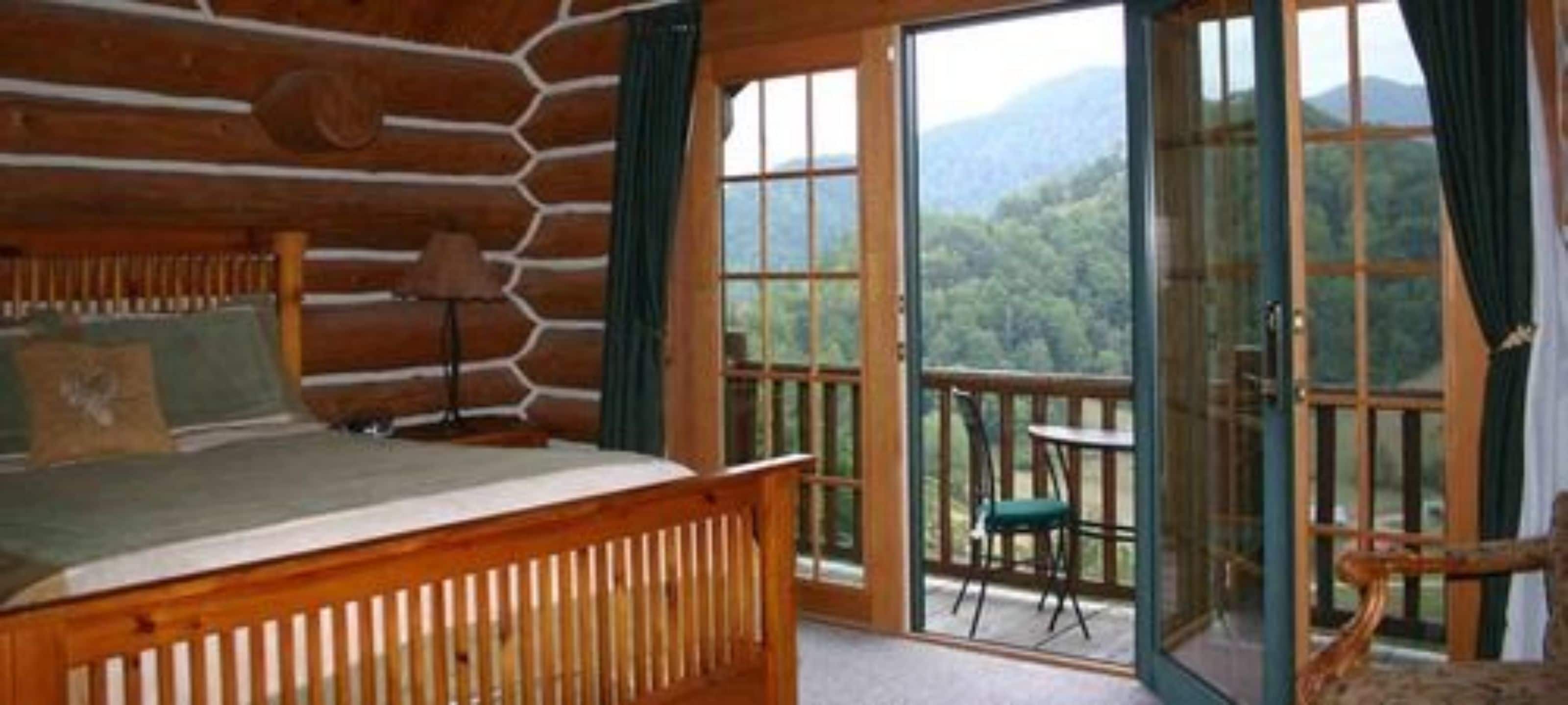 A log cabin bedroom with a bed, a chair, and a table by a sliding glass door leading to a balcony with a mountain view.