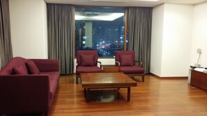 Een moderne woonkamer met een bank, twee fauteuils en een salontafel. Een groot raam biedt uitzicht op een stadsbeeld 's nachts.