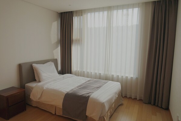 Una cama bien hecha con una manta gris, una mesita de noche de madera y un amplio ventanal con cortinas traslúcidas.
