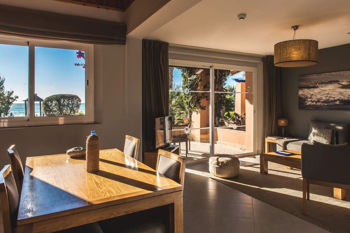 A spacious living area with a wooden dining table, a comfortable sofa, and a large window offering a view of the outdoors.