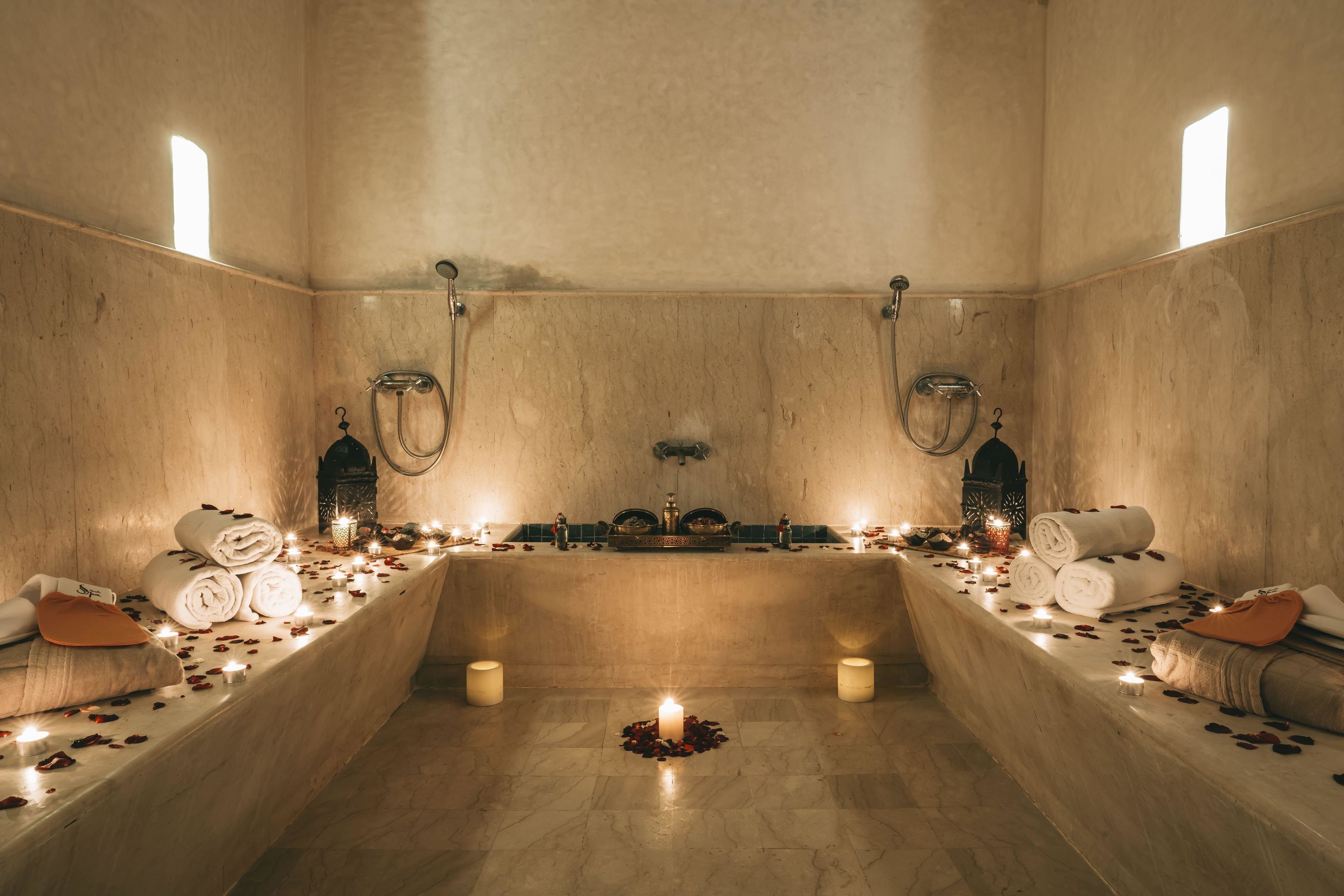 Un cuarto de spa con una bañera central, dos duchas laterales, velas, toallas y pétalos de rosa en el suelo.