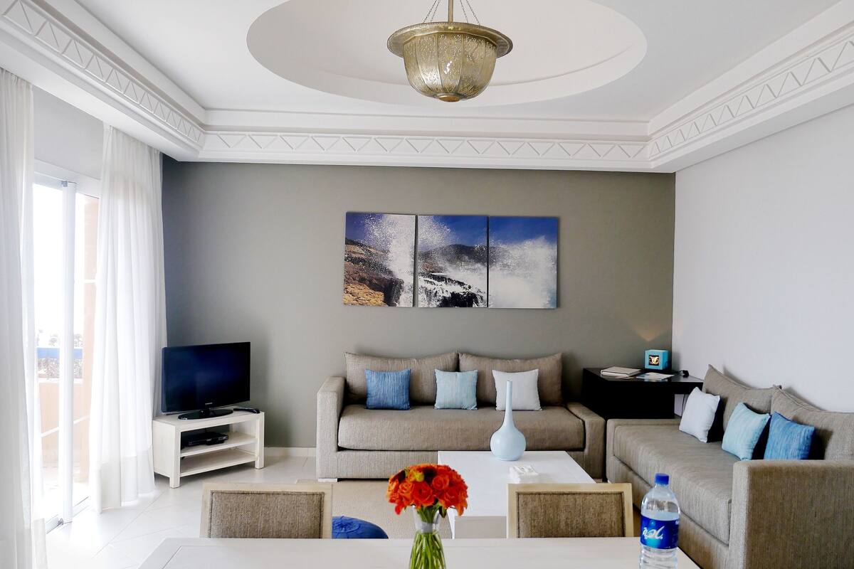 A modern living room with a sofa set, a flat-screen TV, a coffee table with a vase of flowers, and a painting on the wall.