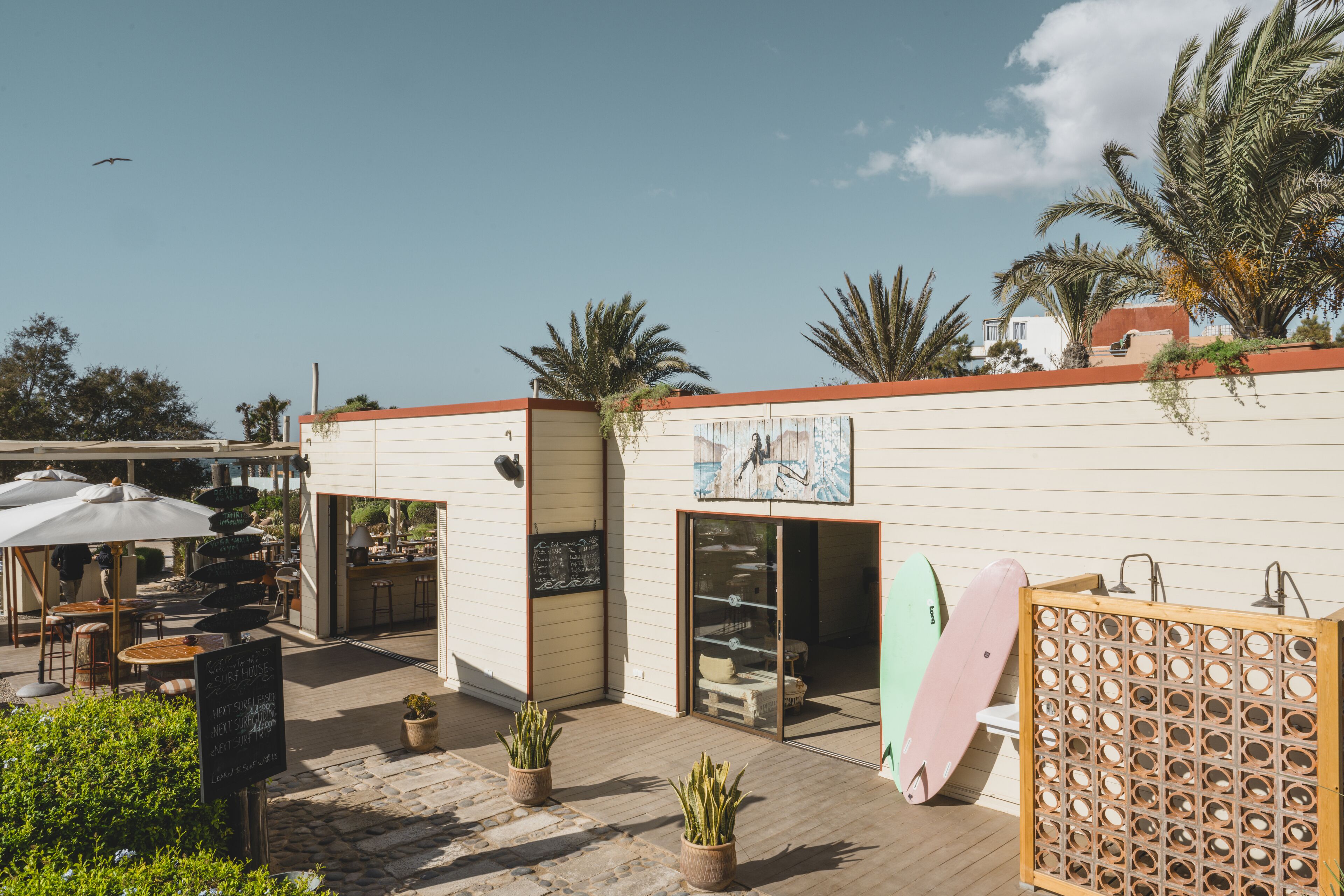 Un espace extérieur aménagé avec des tables et des parasols, un bâtiment avec une enseigne, et des planches de surf appuyées contre le mur.