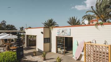 Un espace extérieur aménagé avec des tables et des parasols, un bâtiment avec une enseigne, et des planches de surf appuyées contre le mur.