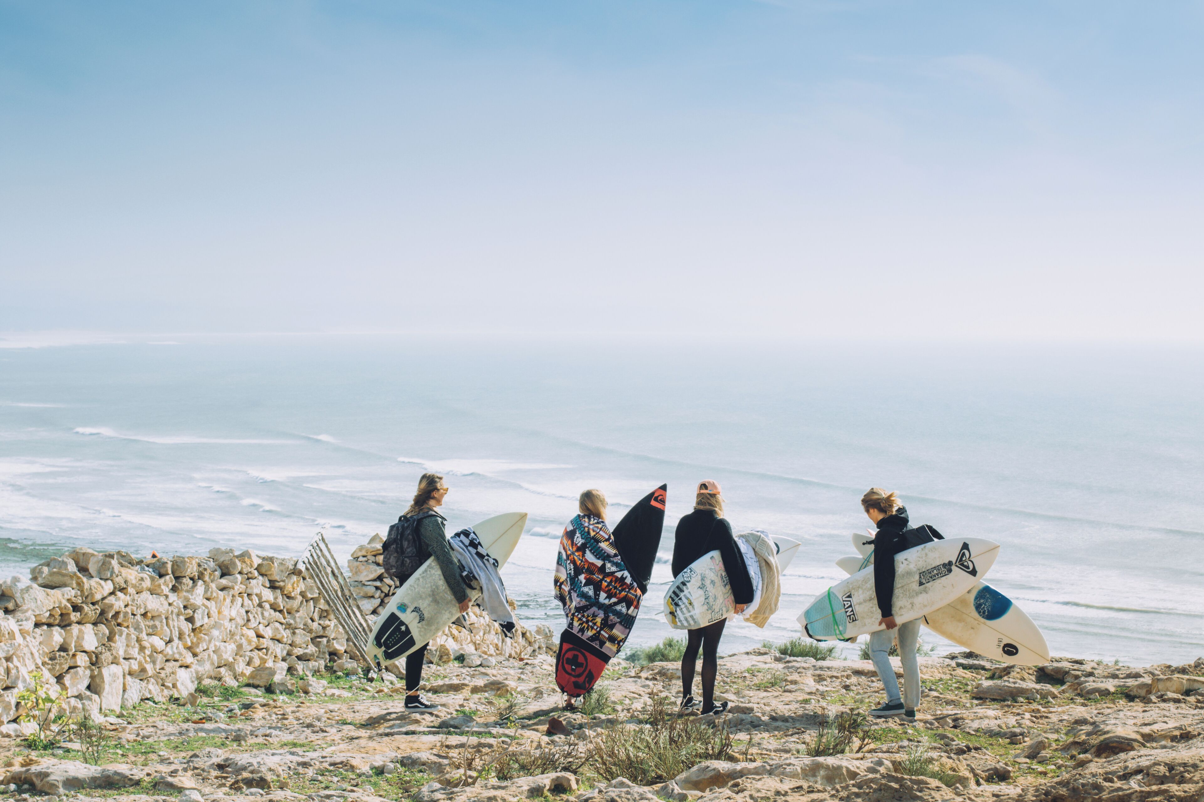 Four surfers with surfboards on a rocky beach with the ocean in the background.
