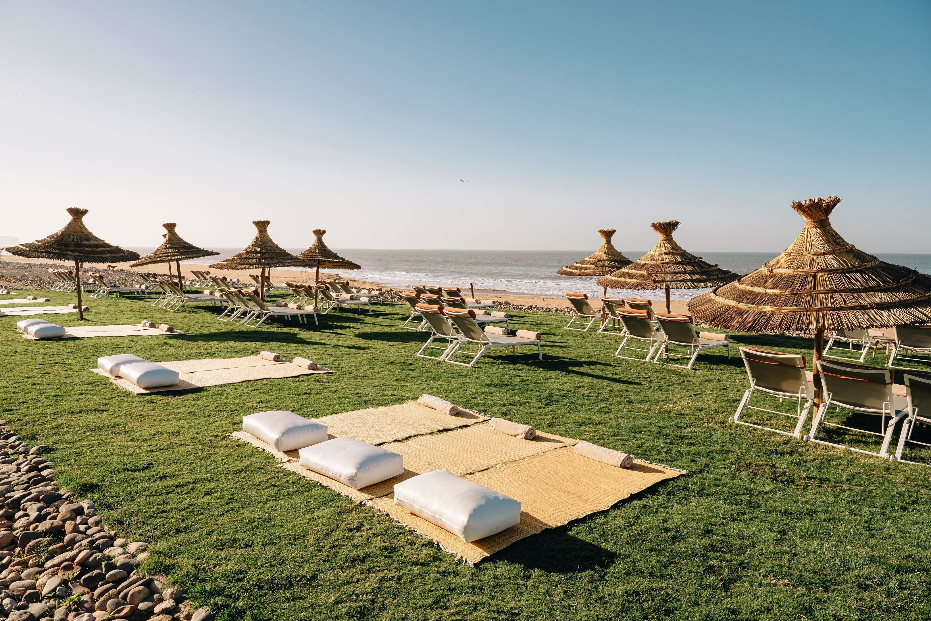 Ein Strandloungebereich mit Reetdachschirmen, Liegestühlen und Sonnenliegen, die auf einer Rasenfläche angeordnet sind.