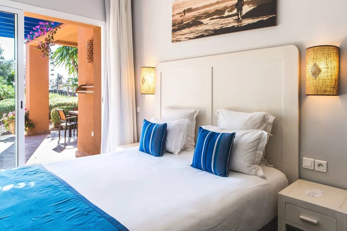 A hotel room with a bed, two pillows, a nightstand, and a wall-mounted lamp.