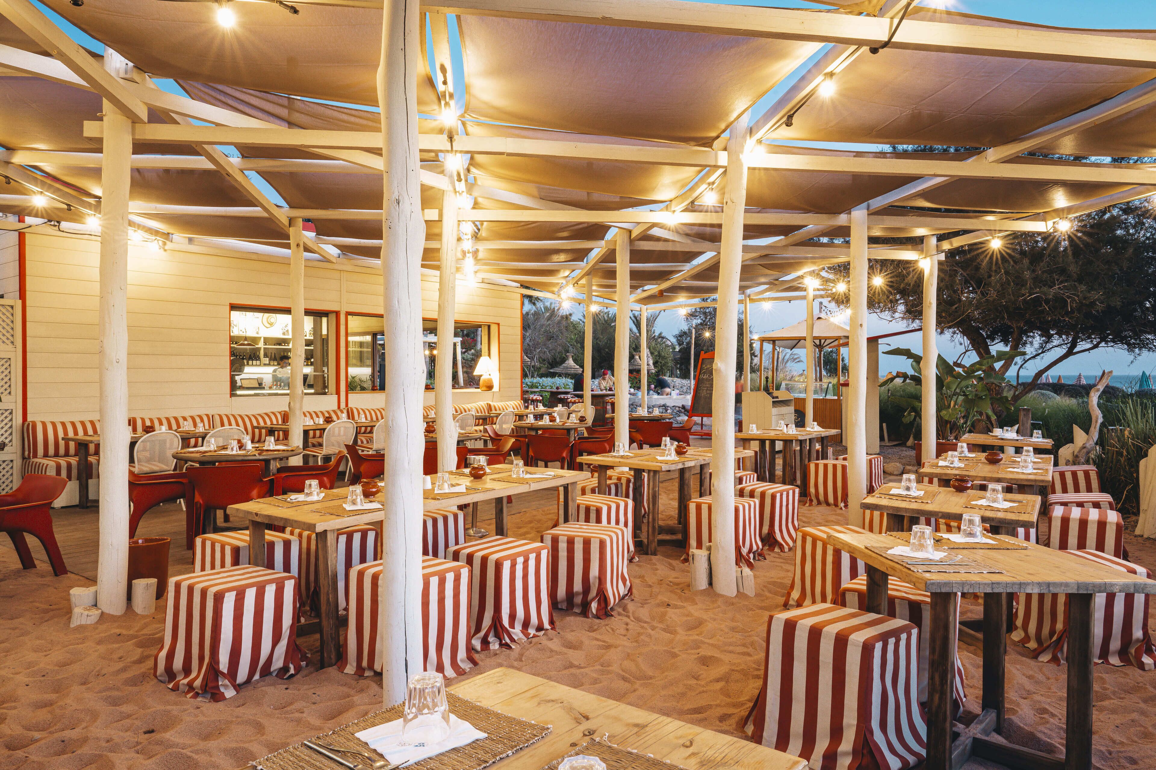 An outdoor dining area with striped tables and chairs, covered by a canopy, and wooden pillars.