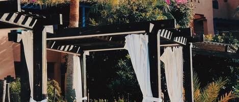 Un lit à baldaquin en bois, surmonté de rideaux blancs, se dressant sur un fond de palmiers et d’un bâtiment à la façade beige.