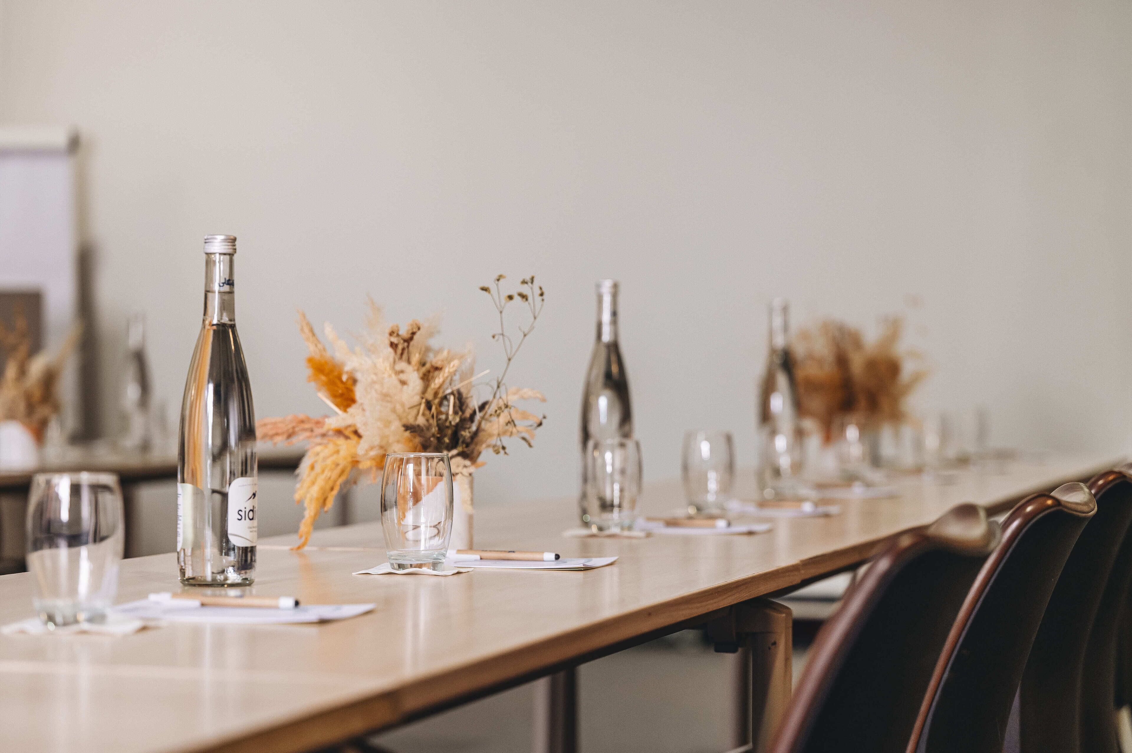 Une longue table à manger, dressée avec des bouteilles de vin, des verres et un centre de table composé de fleurs séchées.