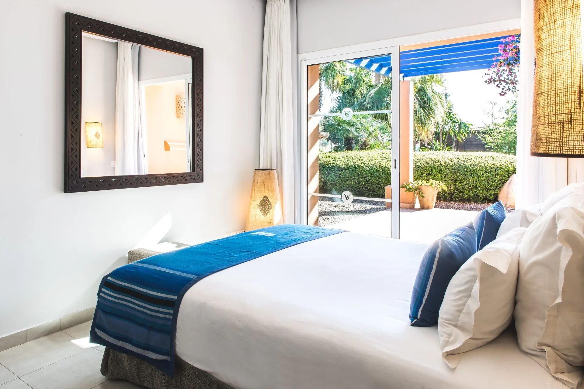 A hotel room with a bed, a large mirror, a nightstand, and a view of a garden through a sliding glass door.