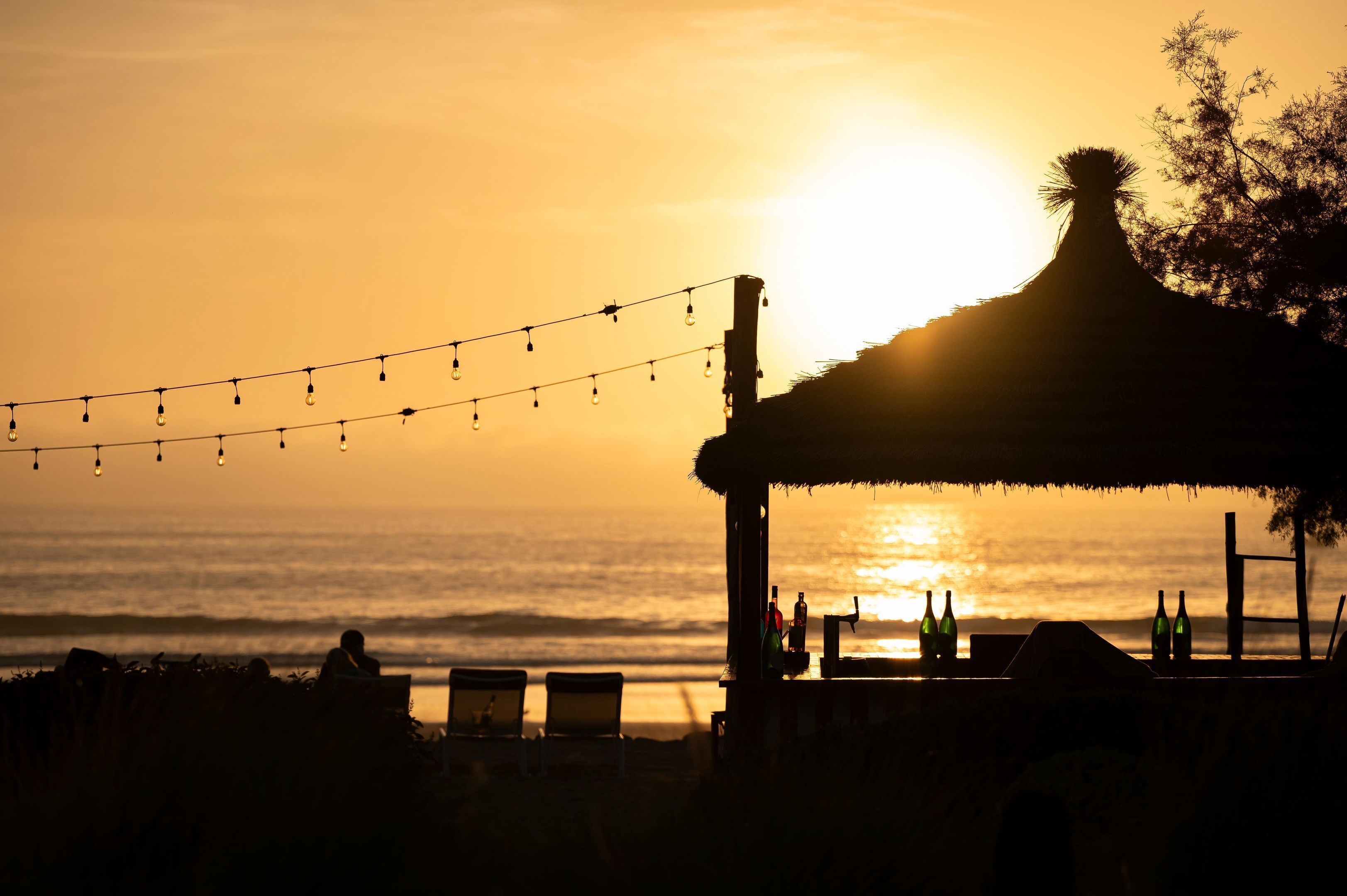A beach scene with a thatched-roof structure, string lights, and bottles on a table.