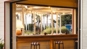 Zona de bar al aire libre con taburetes de madera, una mesa y vistas a palmeras y un barco azul al fondo.