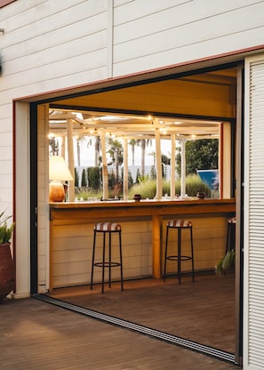 An outdoor bar area with wooden stools, a table, and a view of palm trees and a blue boat in the background. - Paradis Plage Surf Yoga & Spa resort (Tamri)