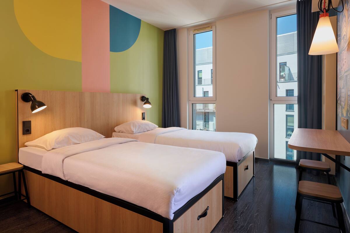 A hotel room with two beds, a desk, and large windows.
