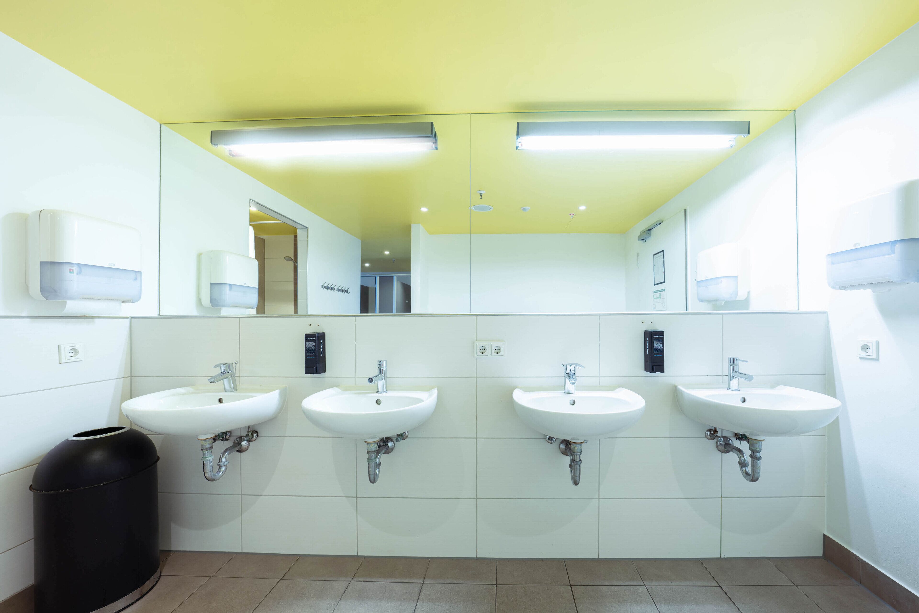 Ein öffentliches WC mit vier Waschbecken, einem Mülleimer und Handdesinfenspender.