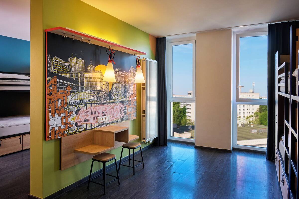 Habitació moderna amb un mural de paisatge urbà, un escriptori de fusta, dos tamborets i grans finestres que donen vista a edificis.