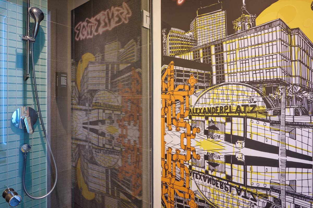 Un bany amb dutxa, paret amb rajoles i un mural d'un paisatge urbà.
