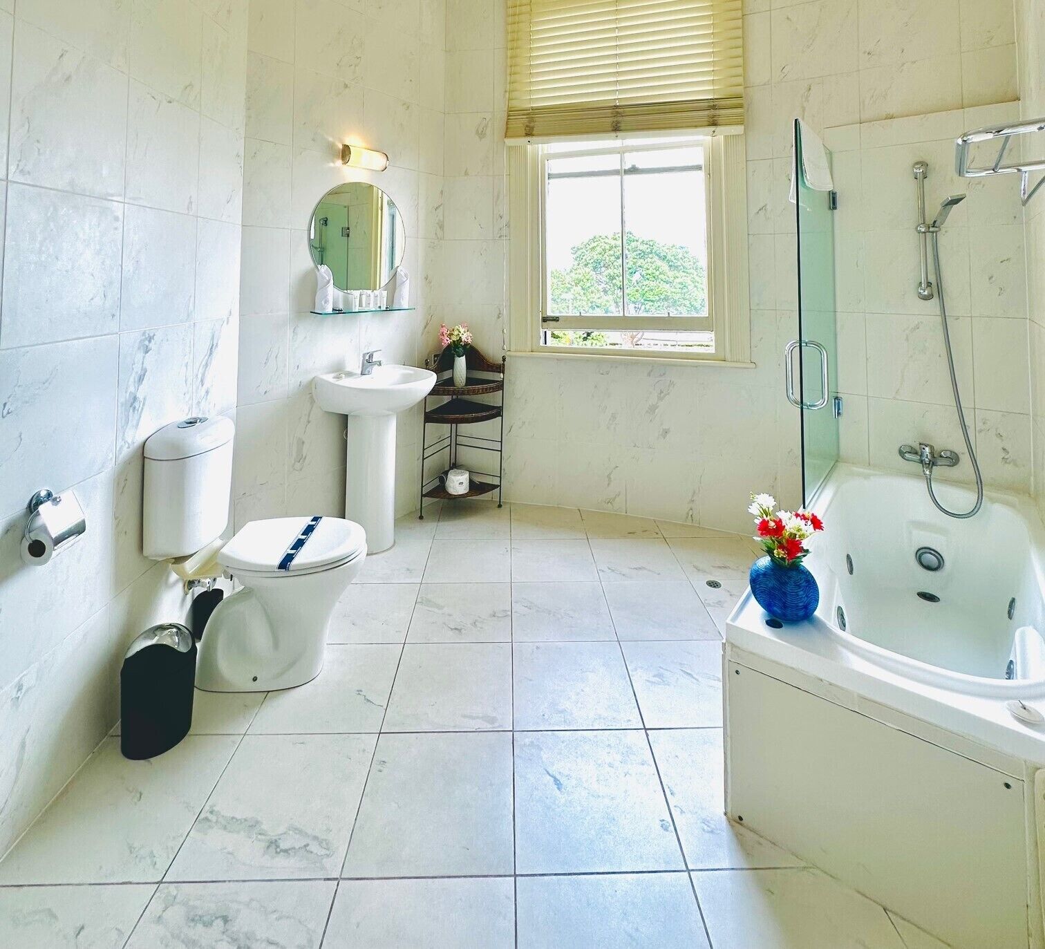 A bathroom with a toilet, sink, and bathtub. There is a window with blinds, a mirror, and a shower.