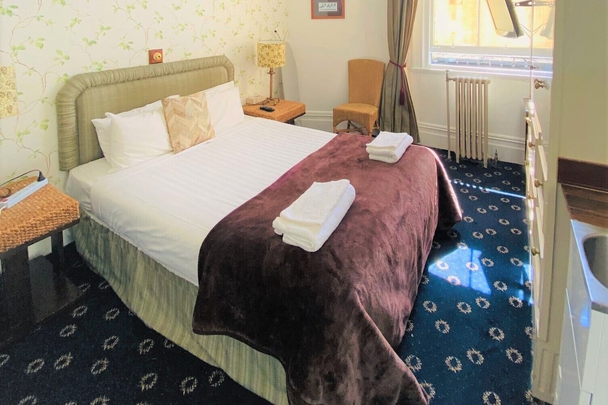 A hotel room with a large bed, a chair, a desk, a window with curtains, and a radiator.