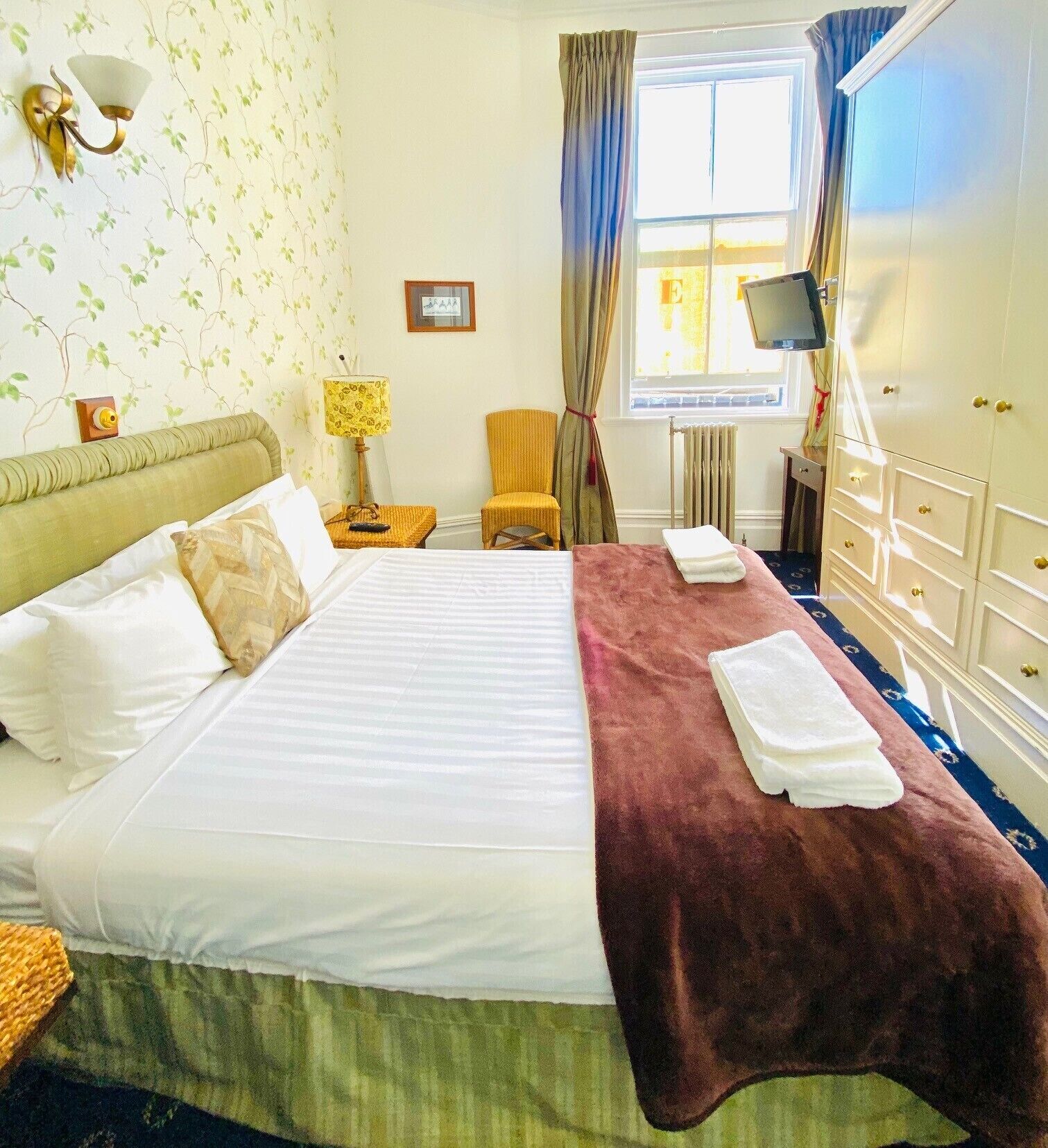 A hotel room with a large bed, a bedside table with a lamp, a chair, a TV, and a wardrobe.