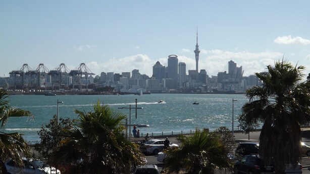 A city skyline with a prominent tower, a waterfront with boats, and a parking area with cars and palm trees. - The Esplanade Hotel (Auckland)