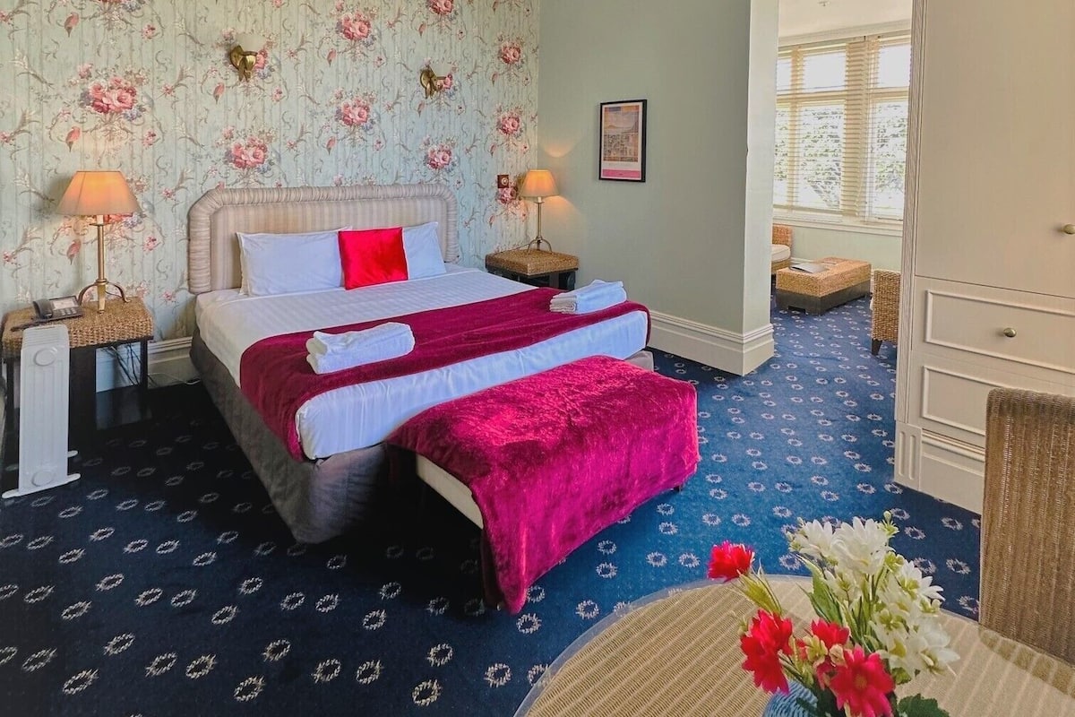 A hotel room with a bed, two bedside tables, a dresser, and a window with blinds.