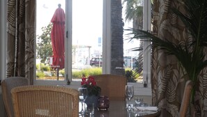 A hotel room with a large window, wicker furniture, a table set with a vase of flowers, and a red umbrella outside. - The Esplanade Hotel (Auckland)