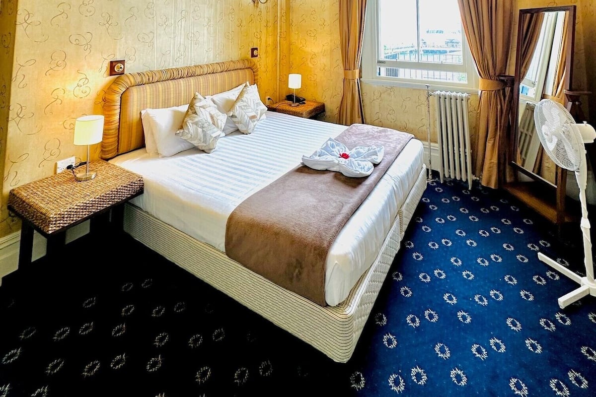 A hotel room with a large bed, a bedside table with a lamp, a standing fan, a window with curtains, and a view of the cityscape.