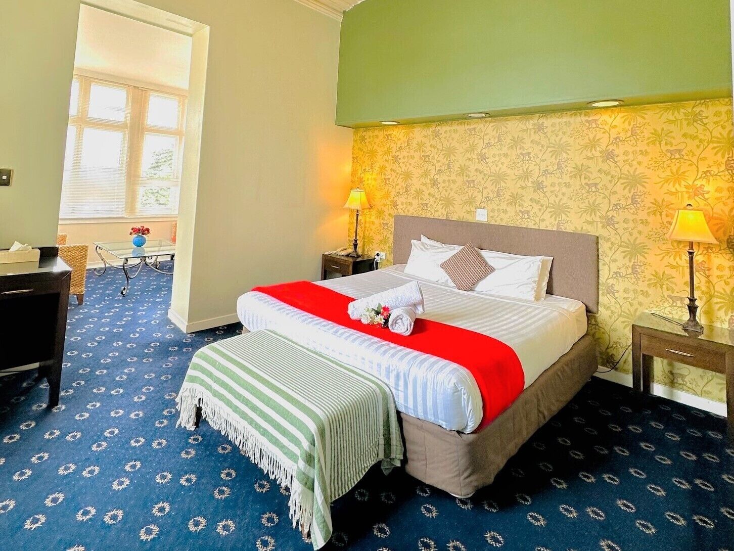 A hotel room with a large bed, two bedside lamps, a window with blinds, a desk, and a chair.