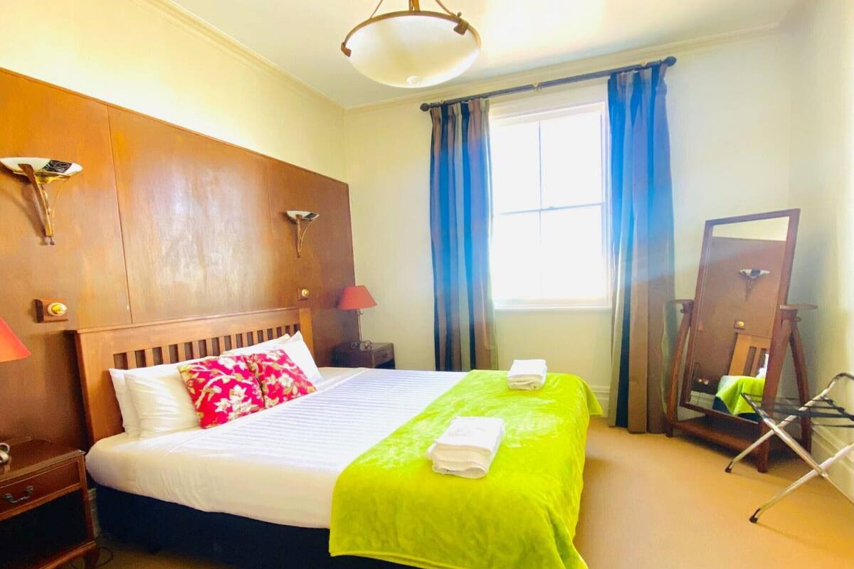 A hotel room with a wooden headboard, a bed with white and green bedding, a nightstand with a lamp, a window with blue curtains, and a mirror on the wall.