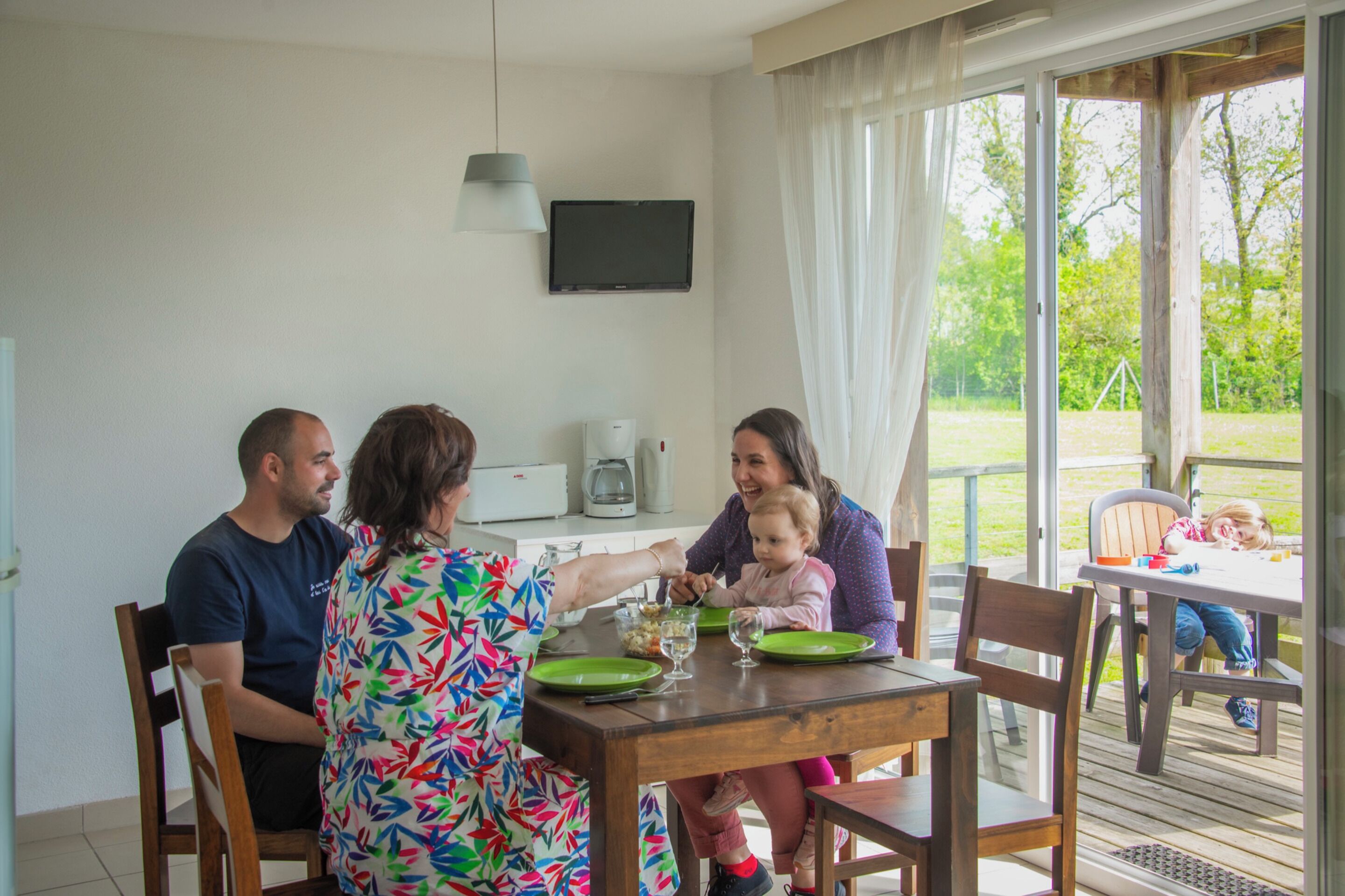 A family dining at a wooden table with a TV mounted on the wall and a view of the outdoors.