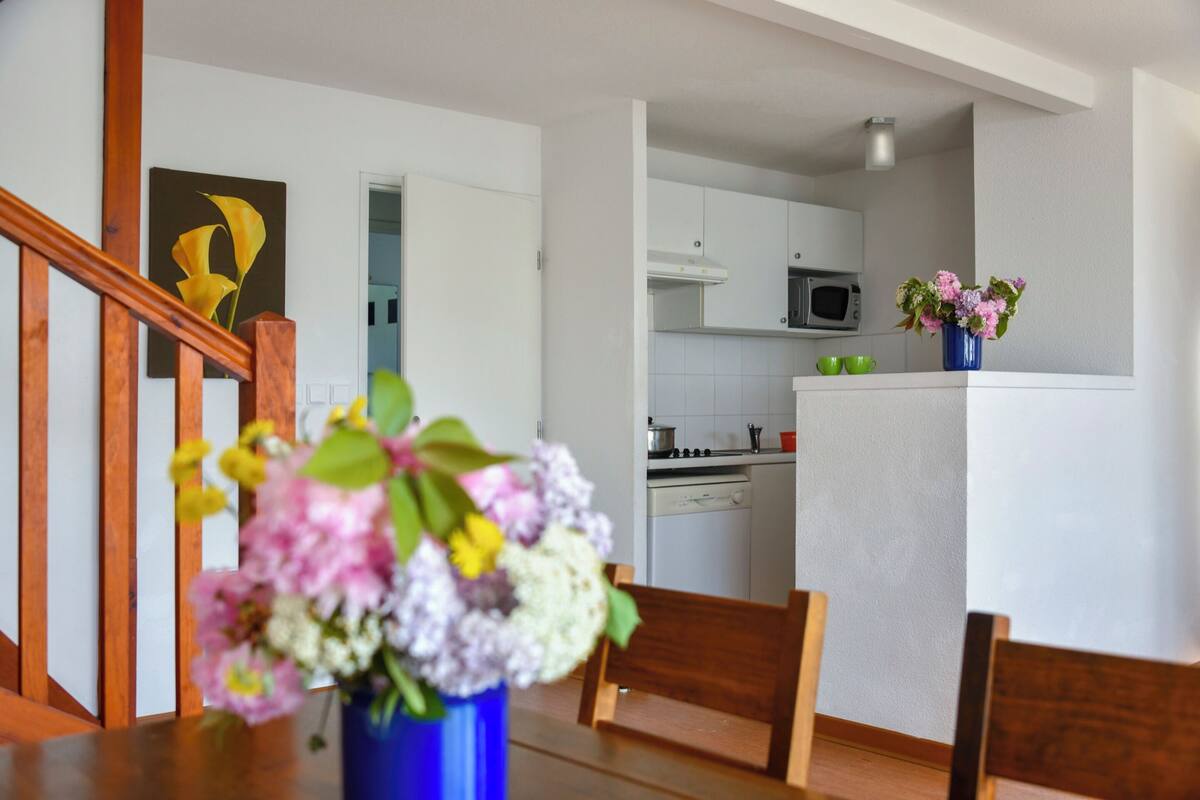 Een eetgedeelte met een houten tafel en stoelen, een vaas met bloemen, en een keuken op de achtergrond met witte kasten en een magnetron.
