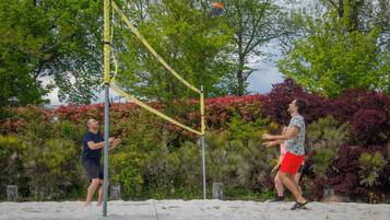 Due persone giocano con una rete da pallavolo in un'area sabbiosa, con alberi e cespugli sullo sfondo.