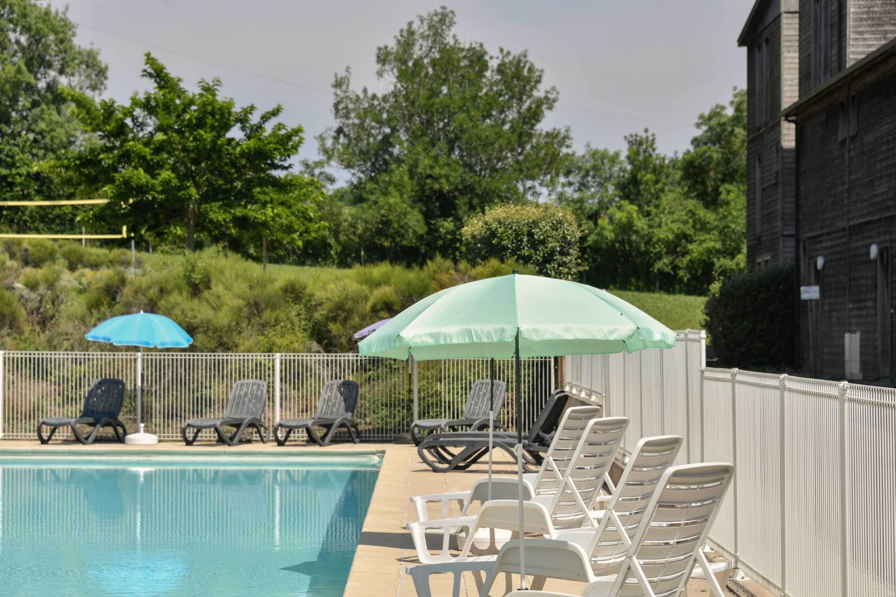 A swimming pool with lounge chairs and umbrellas, surrounded by a fence and greenery.