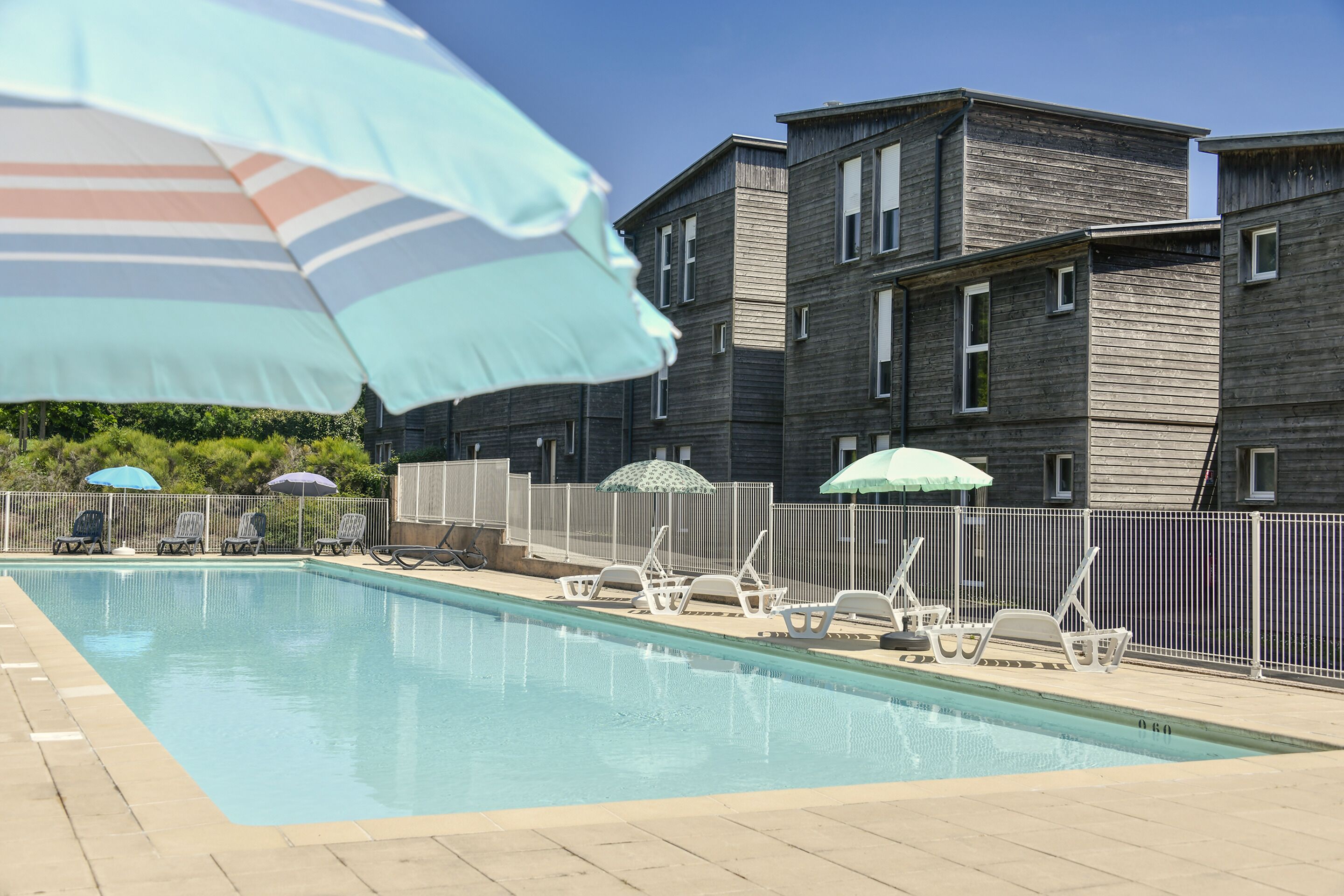 A swimming pool with lounge chairs and umbrellas, surrounded by a fence and a building with multiple windows.